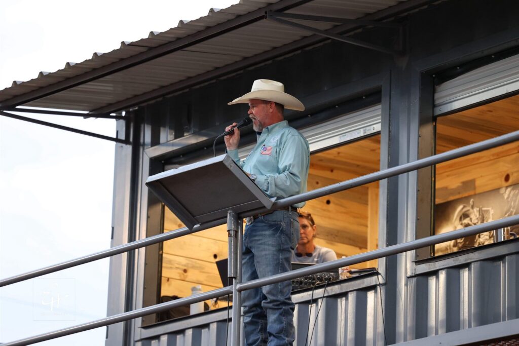 JR McLelland announcing from an elevated announcer’s stand, speaking into a microphone at the railing.