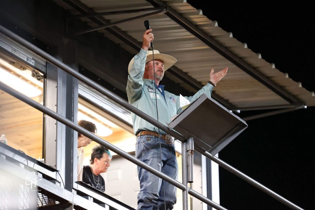 JR McLelland on the announcer’s stand at night, raising a microphone with his other arm outstretched toward the arena.