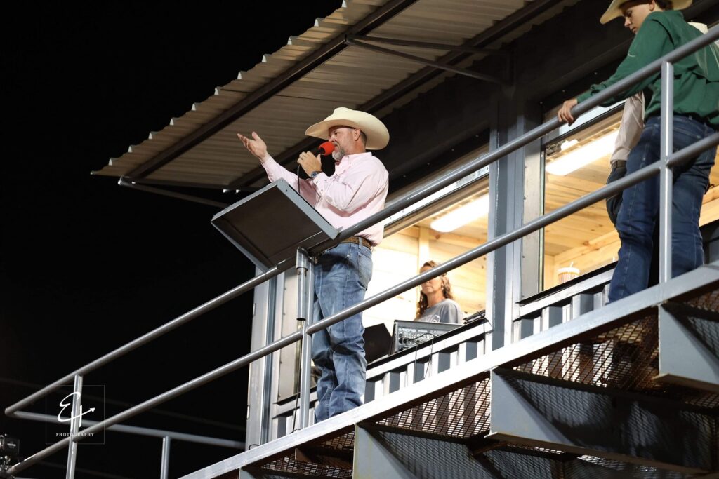 JR McLelland on the announcer’s stand at night in a pink shirt, gesturing toward the arena while speaking into a red microphone.
