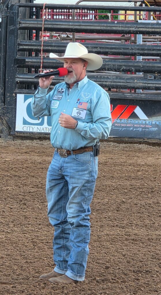 JR McLelland standing in a dirt arena in a light blue shirt and white hat, speaking into a red microphone in front of the bucking chutes.