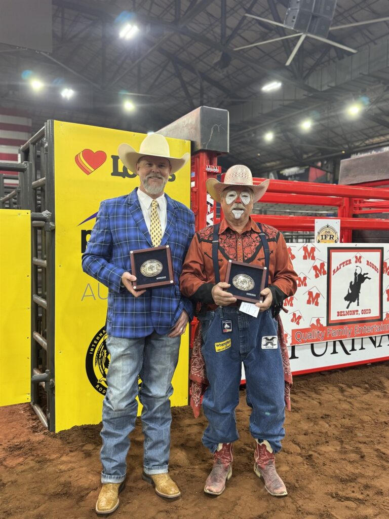 JR McLelland in a blue plaid jacket holding a buckle in a display case, standing beside a rodeo clown holding a matching buckle in an indoor arena.