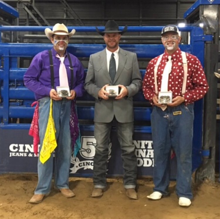 JR McLelland, center in a gray suit and black cowboy hat, holding an award with two other gentlemen in front of blue rodeo equipment.